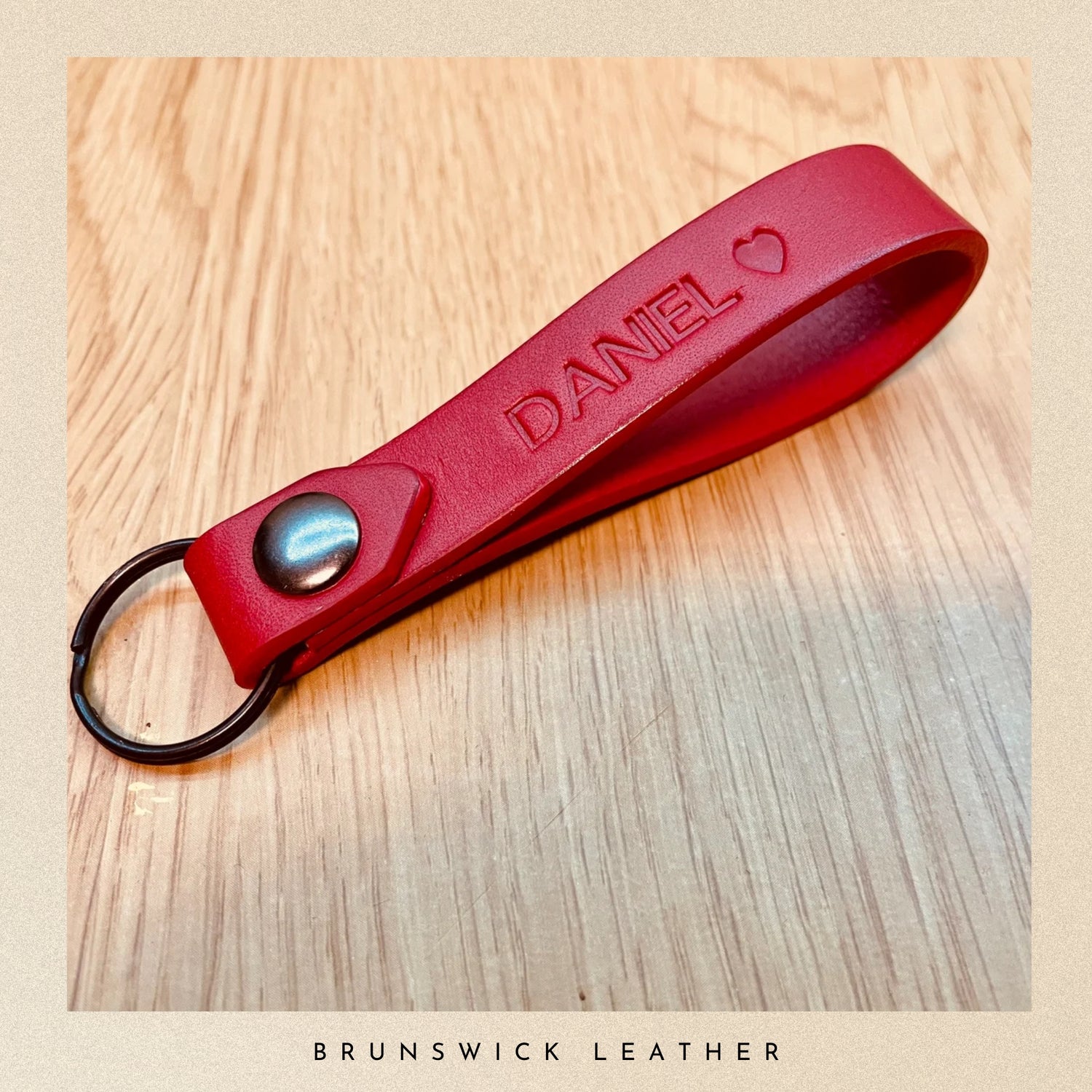 Red leather keychain with 'DANIEL' engraved on a wooden surface, branded 'Brunswick Leather'.