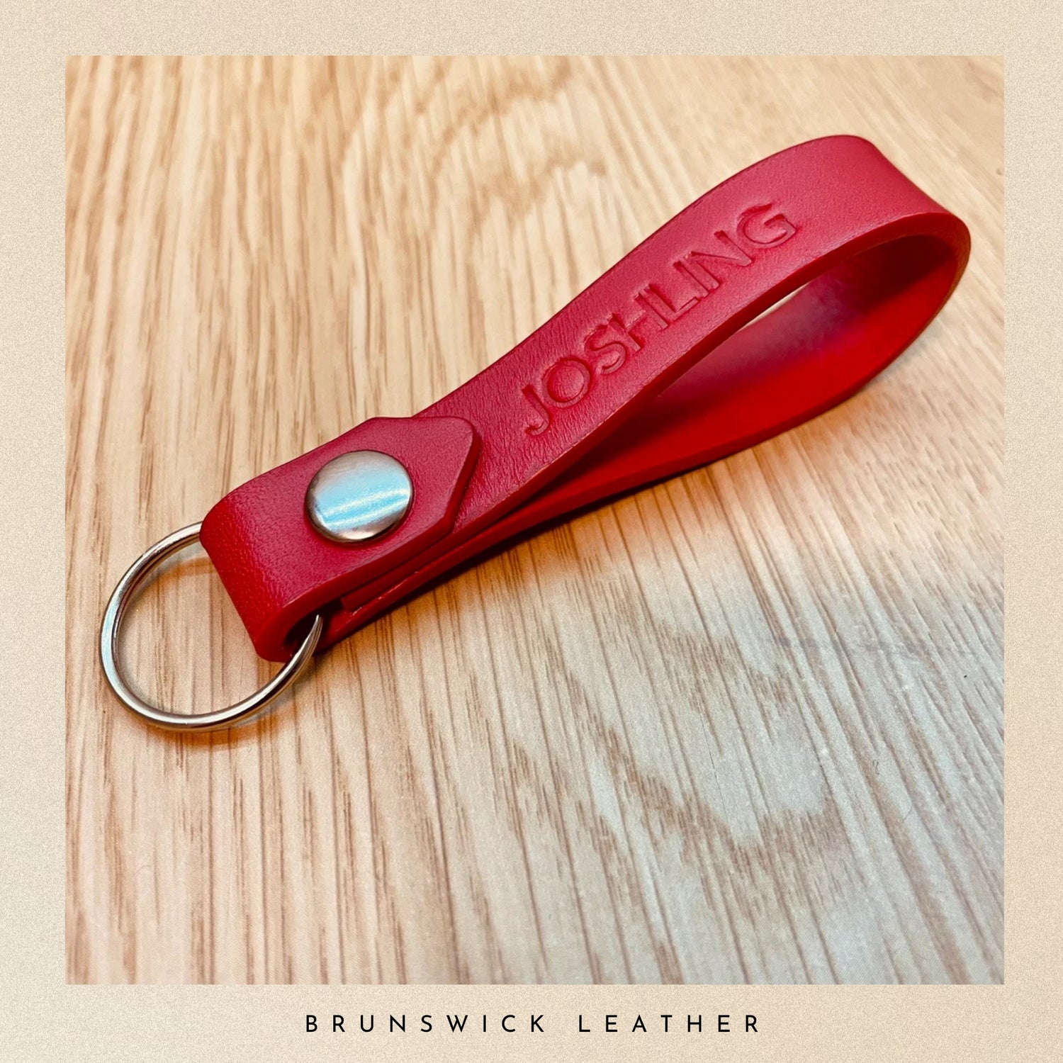 Red leather keychain with 'JOSH-LING' engraving on a wooden surface, branded 'Brunswick Leather'.