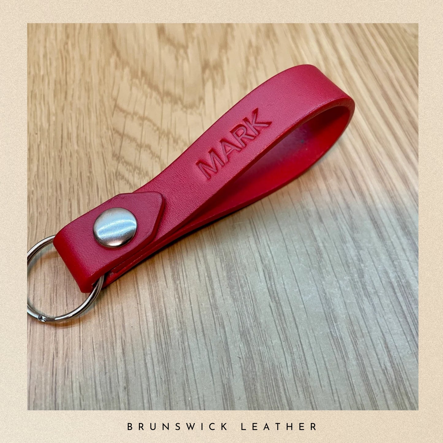 Red leather keychain with 'MARK' engraving on a wooden surface, branded 'Brunswick Leather'.