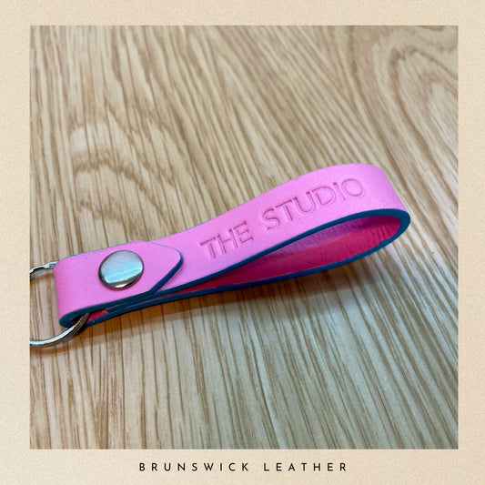 Pink leather keychain with 'THE STUDIO' embossed on a wooden surface, branded 'Brunswick Leather'.