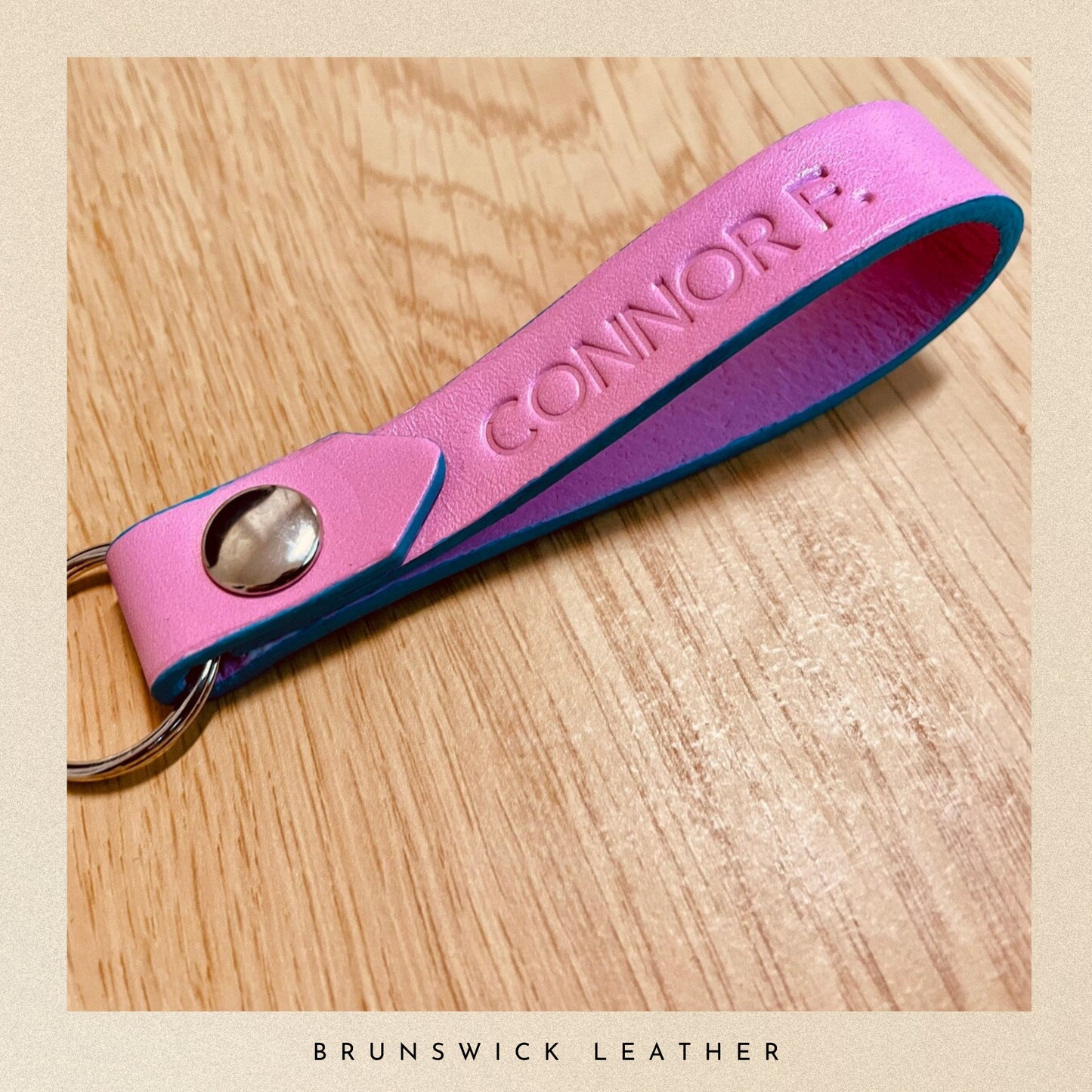 Pink leather keychain with 'CONNOR F.' engraving on a wooden surface, branded 'Brunswick Leather'.