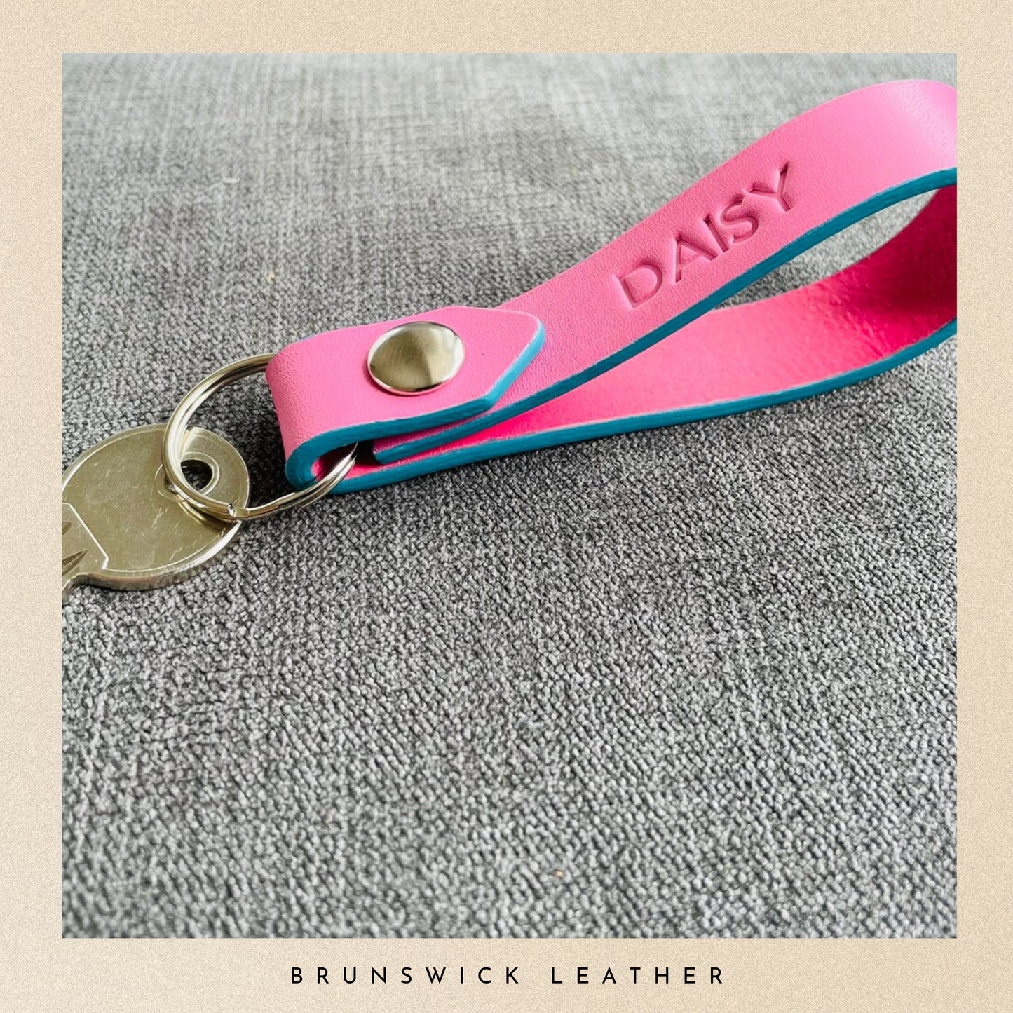 Pink leather key fob with 'DAISY' engraving on a gray surface, branded 'Brunswick Leather'.