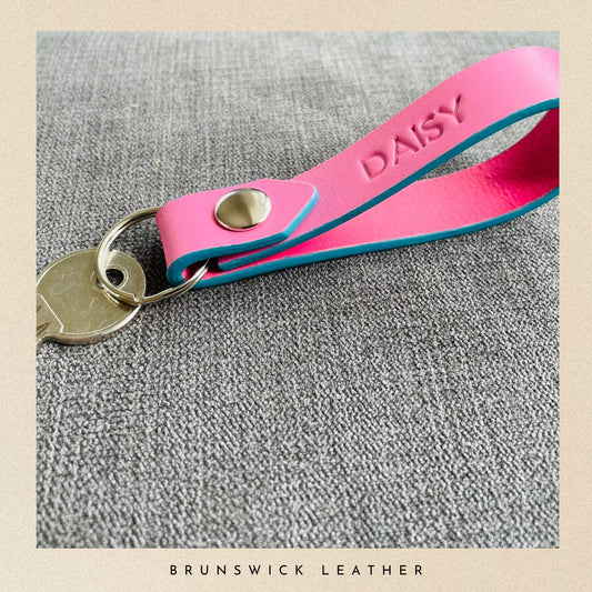 Pink leather key fob with 'DAISY' engraving on a gray surface, branded 'Brunswick Leather'.