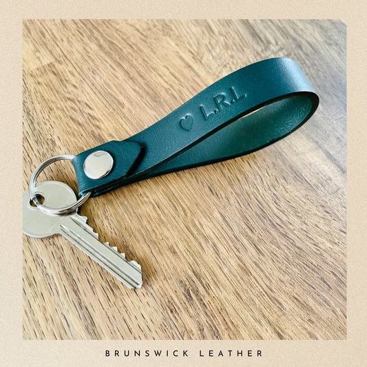 Green leather keychain with engraved initials on a wooden surface, branded 'Brunswick Leather'. 