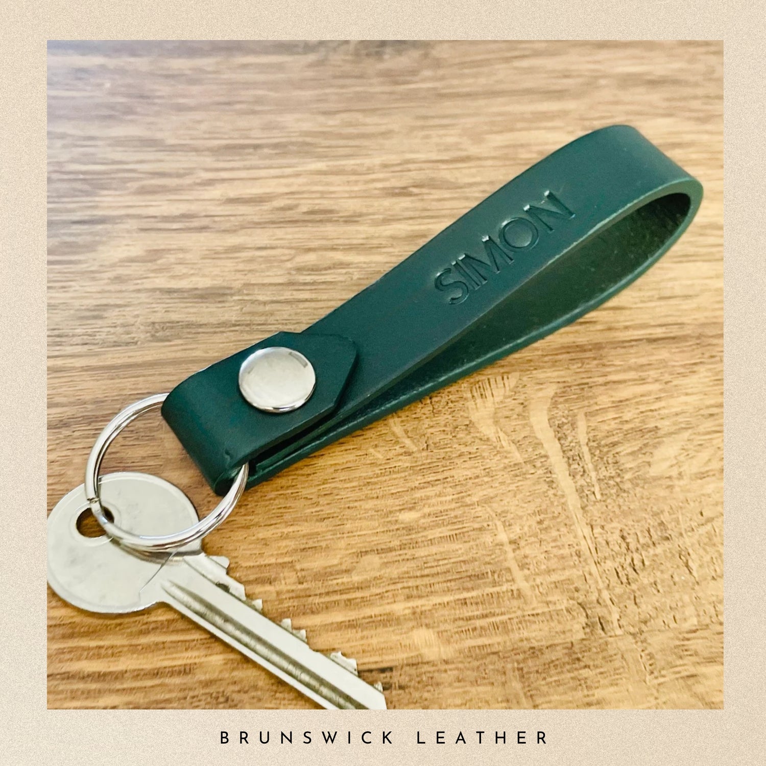 Green leather keychain with 'SIMON' engraving on a wooden surface, branded 'Brunswick Leather'.