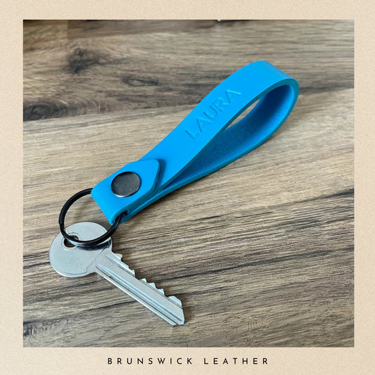 Blue keychain with a key on a wooden surface, branded 'Brunswick Leather'.