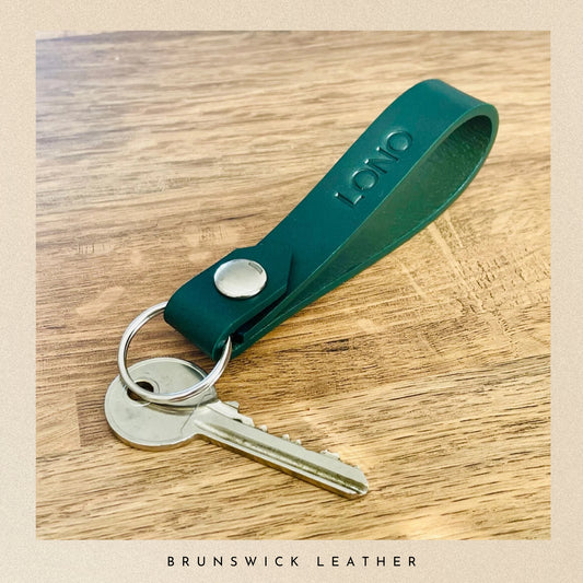 green leather keychain with a key on a wooden surface, branded 'Brunswick Leather'.