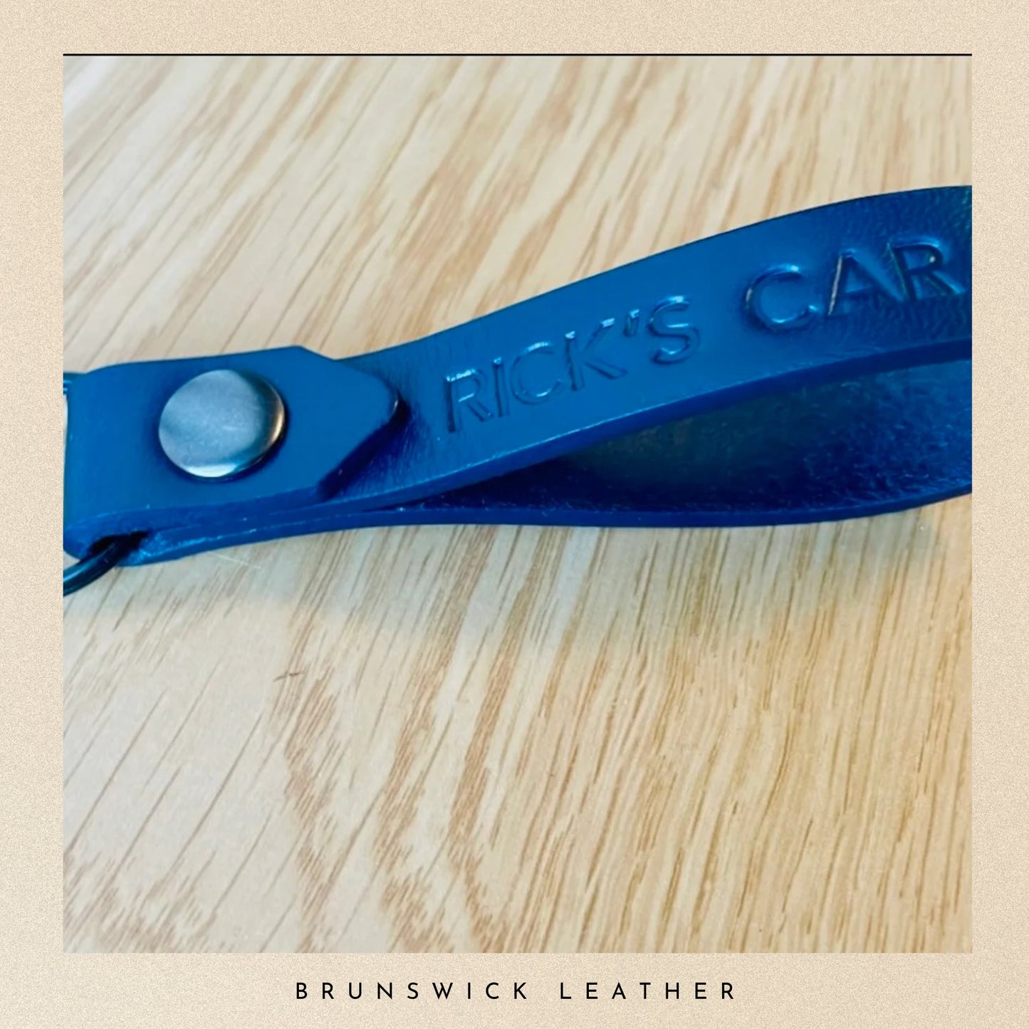 Blue leather car key fob cover with 'Rick's Car' engraving on a wooden surface, branded 'Brunswick Leather'.