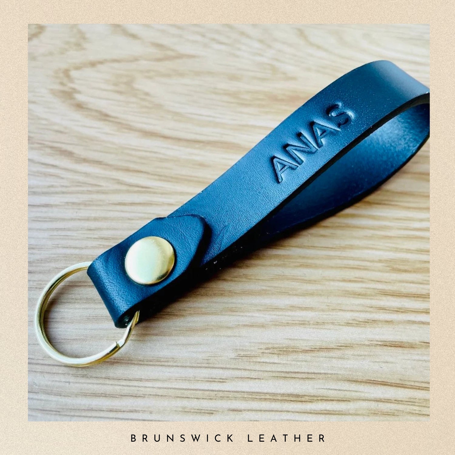Blue leather keychain with 'ANAS' engraving on a wooden surface, branded 'Brunswick Leather'.