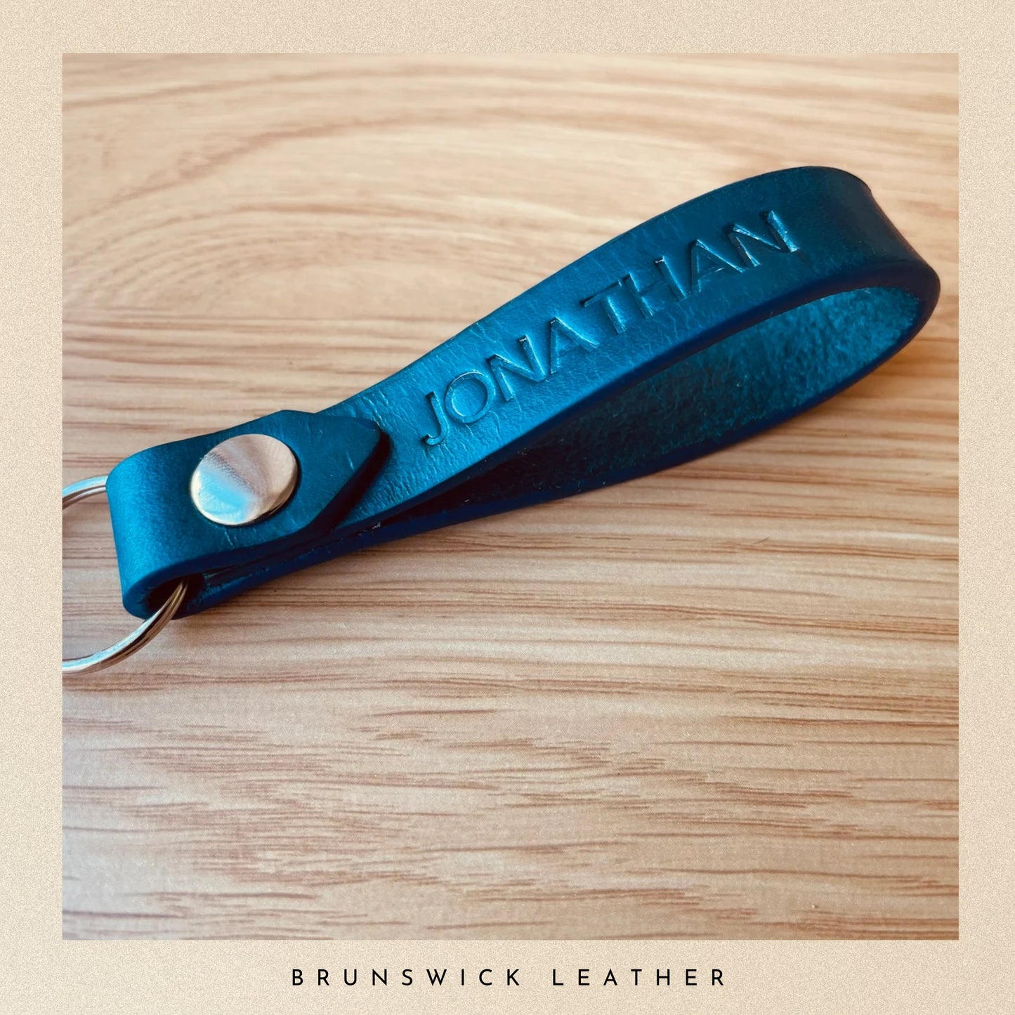Blue leather keychain with 'JONATHAN' engraving on a wooden surface, branded 'Brunswick Leather'.