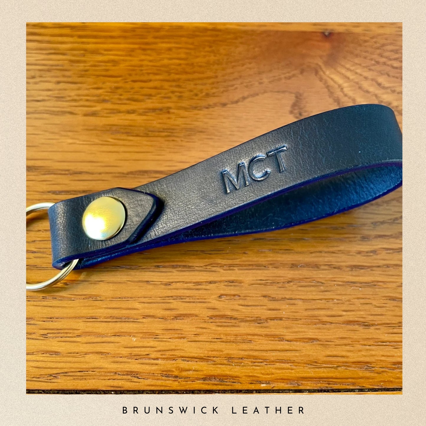Blue leather keychain with 'MCT' engraving on a wooden surface, branded 'Brunswick Leather'.