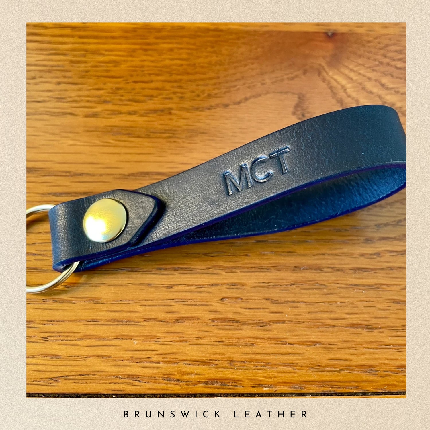Blue leather keychain with 'MCT' engraving on a wooden surface, branded 'Brunswick Leather'.