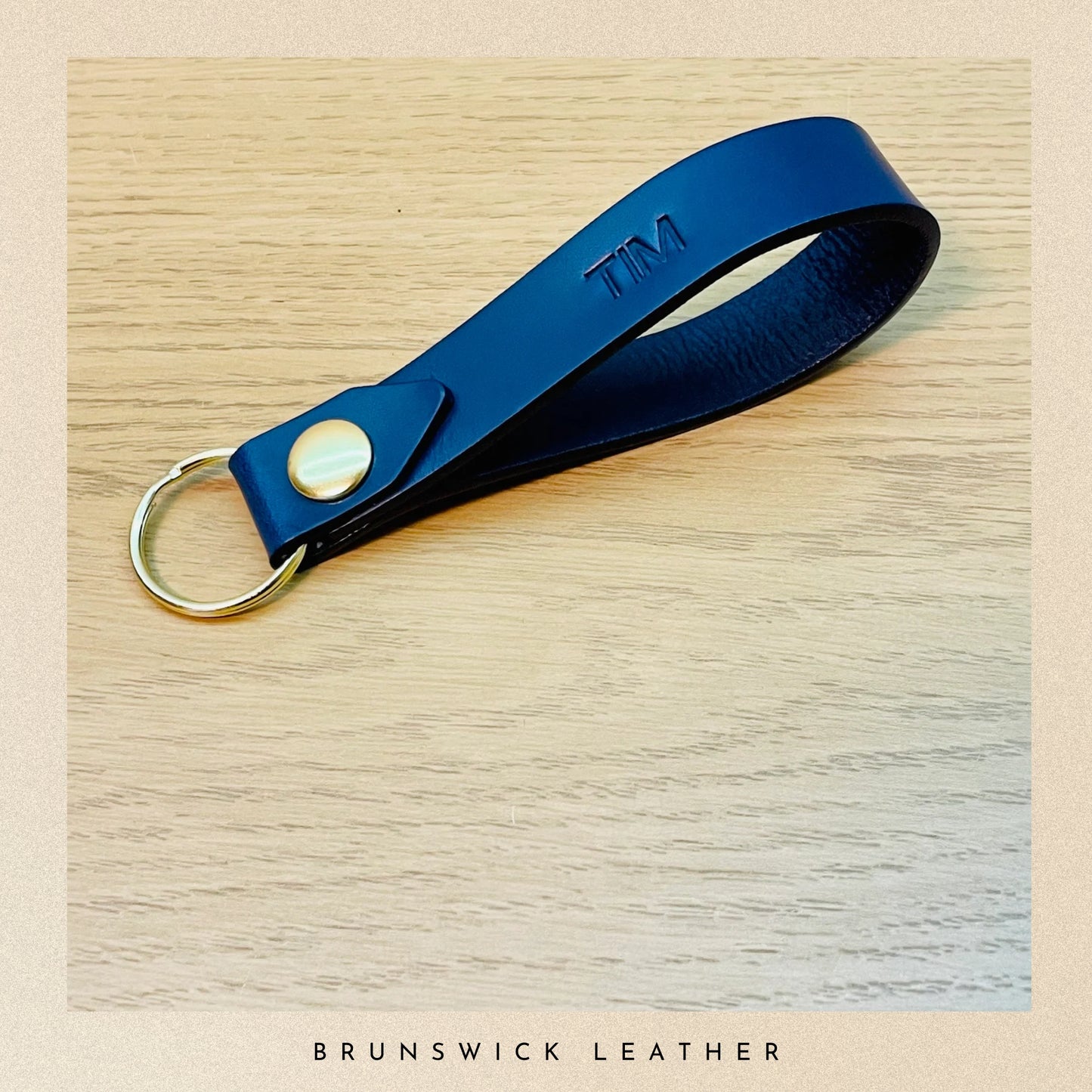 Blue leather keychain with gold ring and 'TIM' engraving on a wooden surface, branded 'Brunswick Leather'.