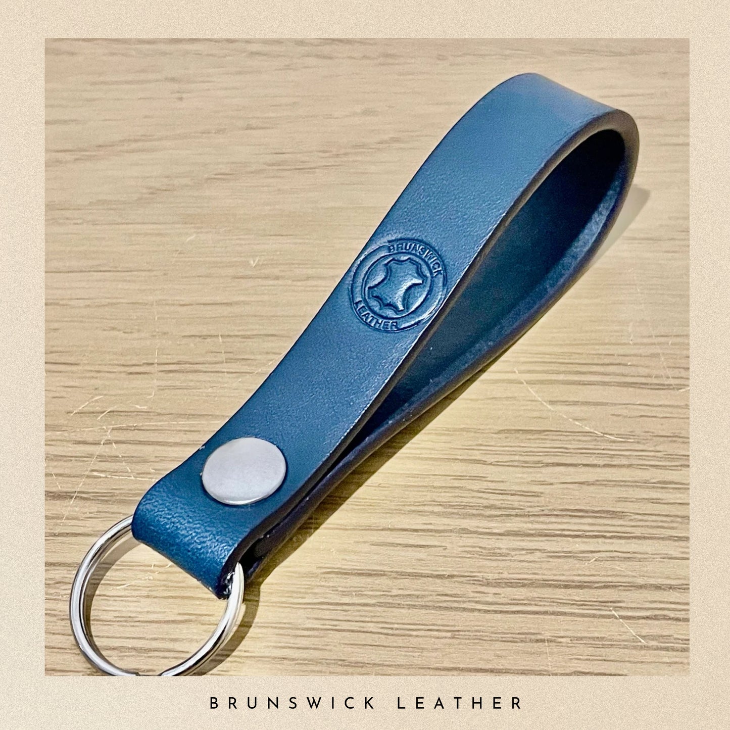 Blue leather keychain with a metal ring on a wooden surface, branded 'Brunswick Leather'.