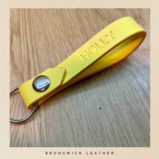 Yellow leather keychain with 'HOLLY' engraved on a wooden surface, branded 'Brunswick Leather'.