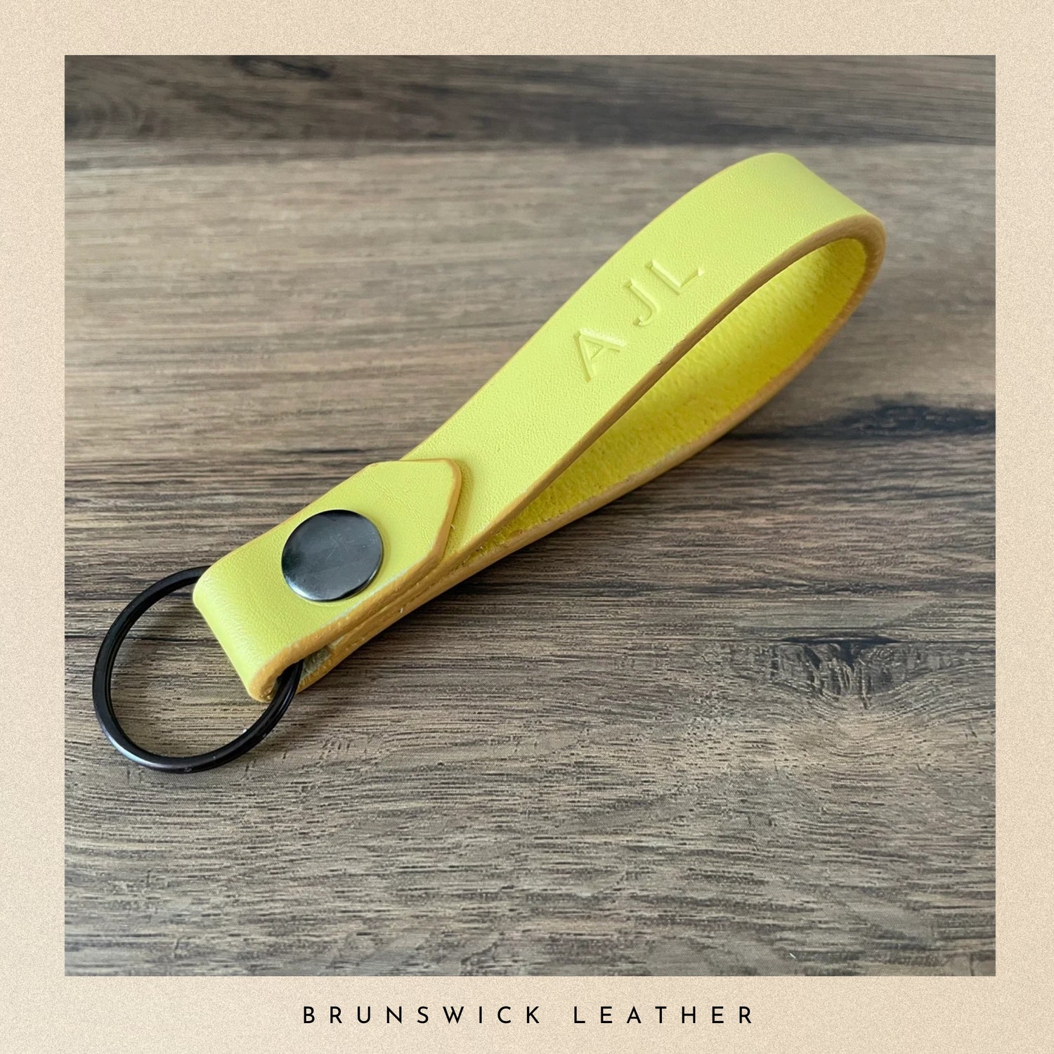 Yellow leather keychain with a metal ring on a wooden surface, branded 'A.J.L'.