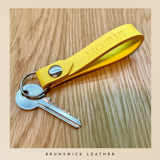 Yellow leather keychain with a key on a wooden surface, branded 'Brunswick Leather'.