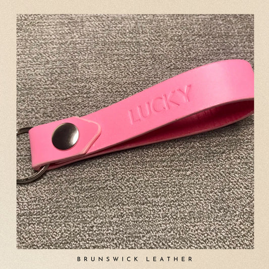Pink leather keychain with 'LUCKY' engraving on a textured surface, branded 'Brunswick Leather'.