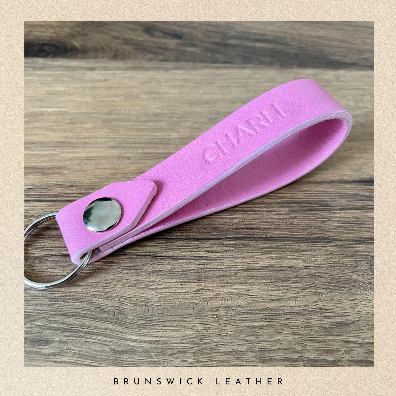 Pink leather keychain with 'CHARM' engraving on a wooden surface, branded 'Brunswick Leather'.