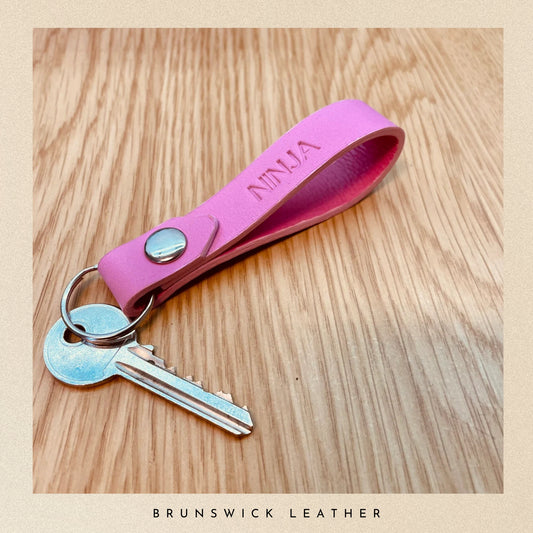 Pink keychain with a key on a wooden surface, branded 'Brunswick Leather'.