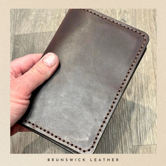 Hand holding a brown leather wallet on a wooden surface with 'Brunswick Leather' branding.