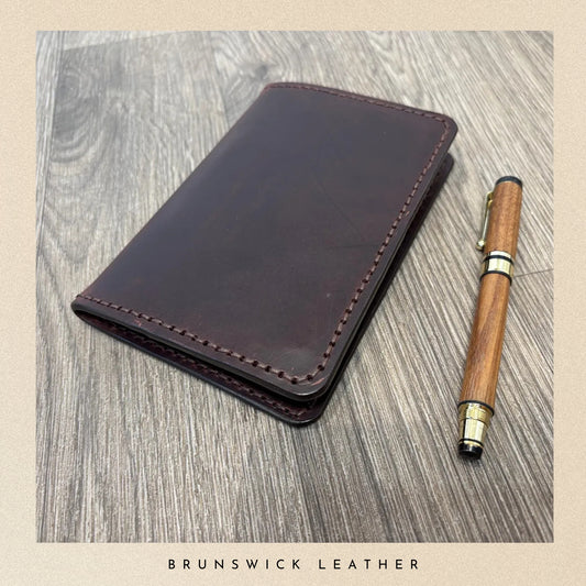 Brown leather wallet and wooden pen on a wooden surface with 'Brunswick Leather' branding.