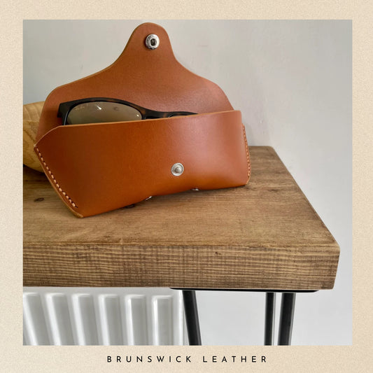 Brown leather eyeglass case with glasses on a wooden surface, branded 'Brunswick Leather'.