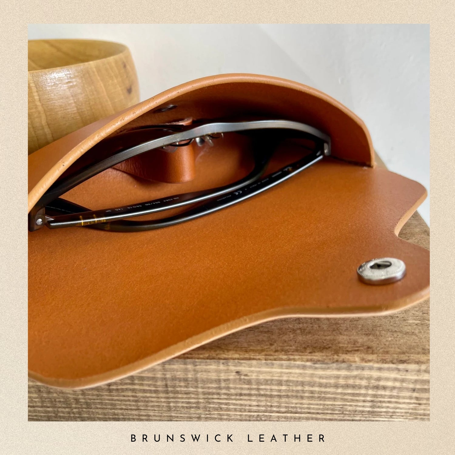 Brown leather case with sunglasses inside, on a wooden surface, branded 'Brunswick Leather'.