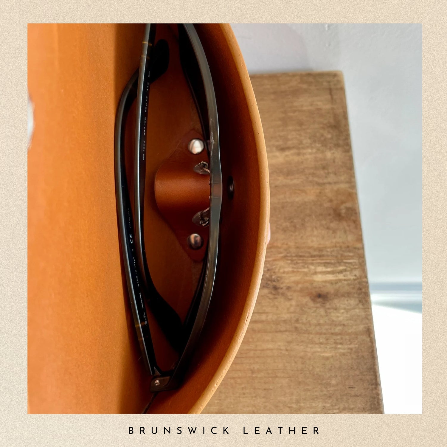Brown leather guitar case with sunglasses on a wooden surface, branded 'Brunswick Leather'.