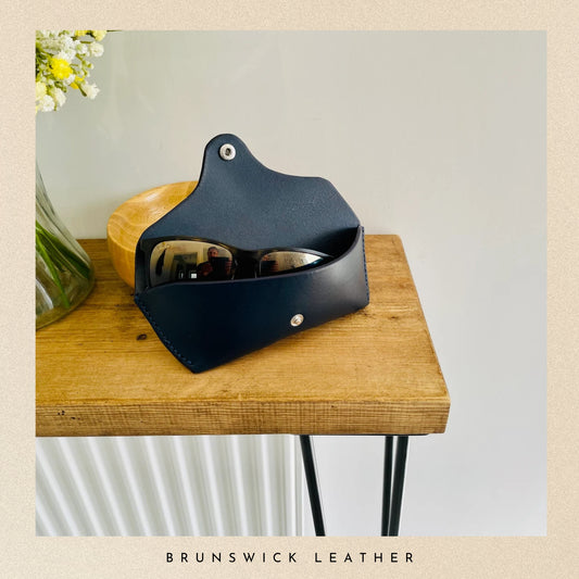 Black leather sunglasses case with sunglasses inside on a wooden surface, branded 'Brunswick Leather'.
