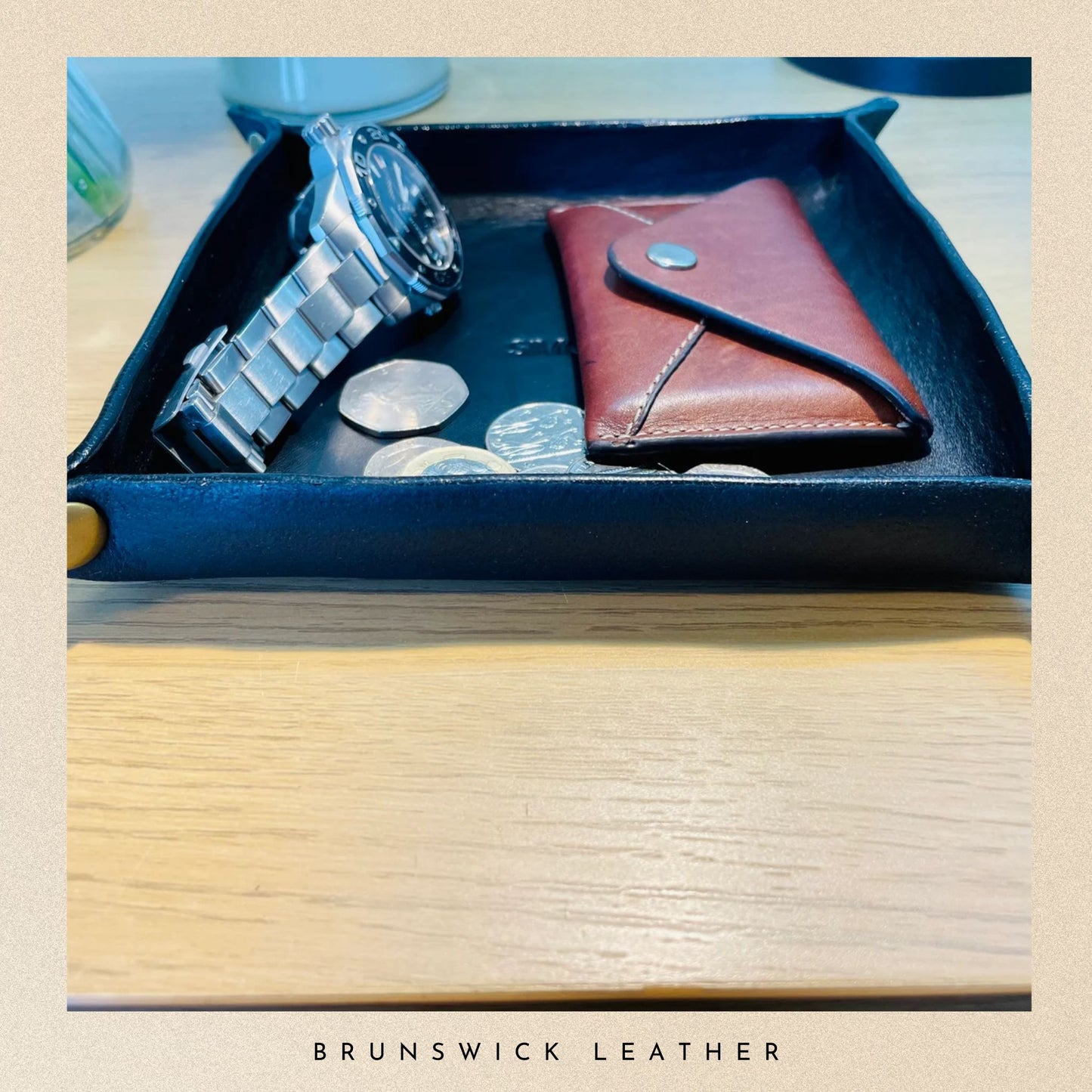 Blue leather tray with a silver watch, brown leather wallet, and coins on a wooden surface. 'Brunswick Leather' branding visible.