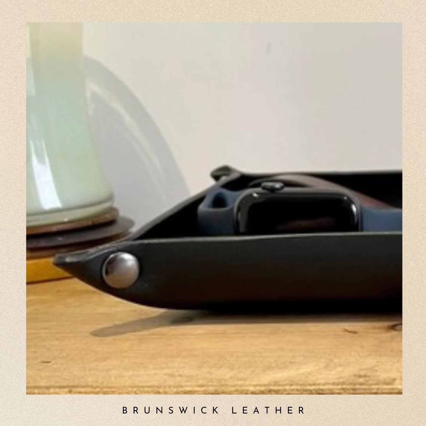 Black leather tray with a silver button on a wooden surface, featuring the brand 'Brunswick Leather'.