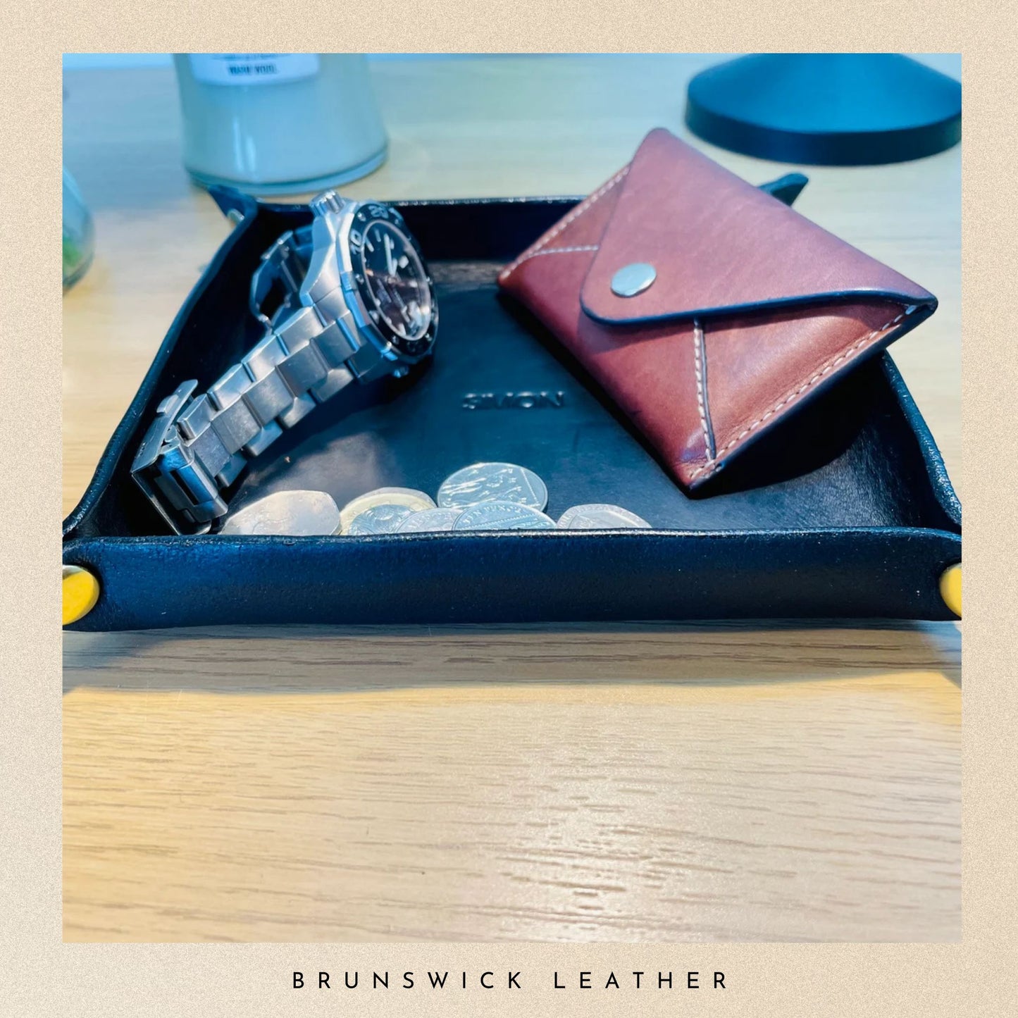 Blue leather tray with a silver watch, red wallet, and coins on a wooden surface with 'Brunswick Leather' branding.