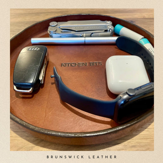 Brown leather tray with various items including car keys, pen, and smartwatch on a wooden surface.