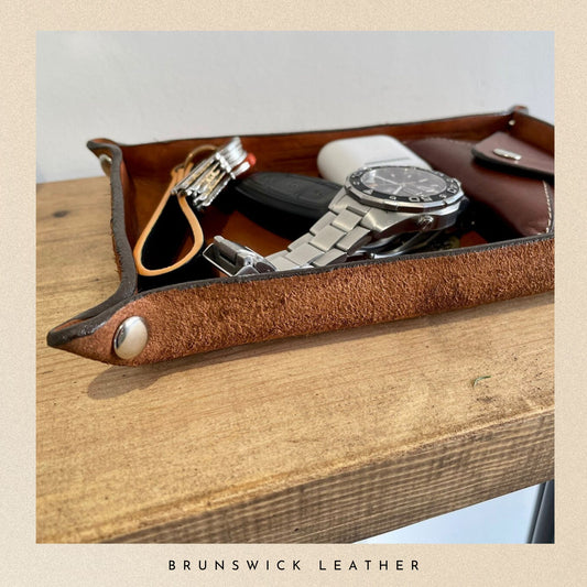 Brown leather tray with watch, keys, and phone on a wooden surface, branded 'Brunswick Leather'.
