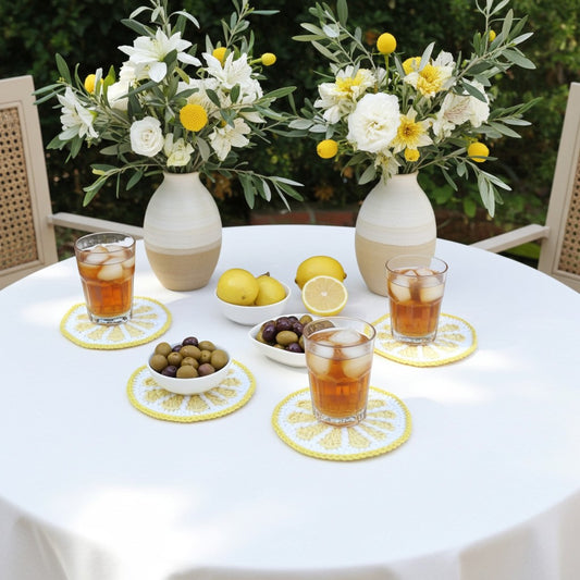 A styled outdoor dining scene with a round white table set with two cream-and-beige ceramic vases filled with white and yellow flowers, lemonade glasses on yellow-and-white lemon-pattern coasters, olives in small white bowls, and lemons, all against a garden background.
