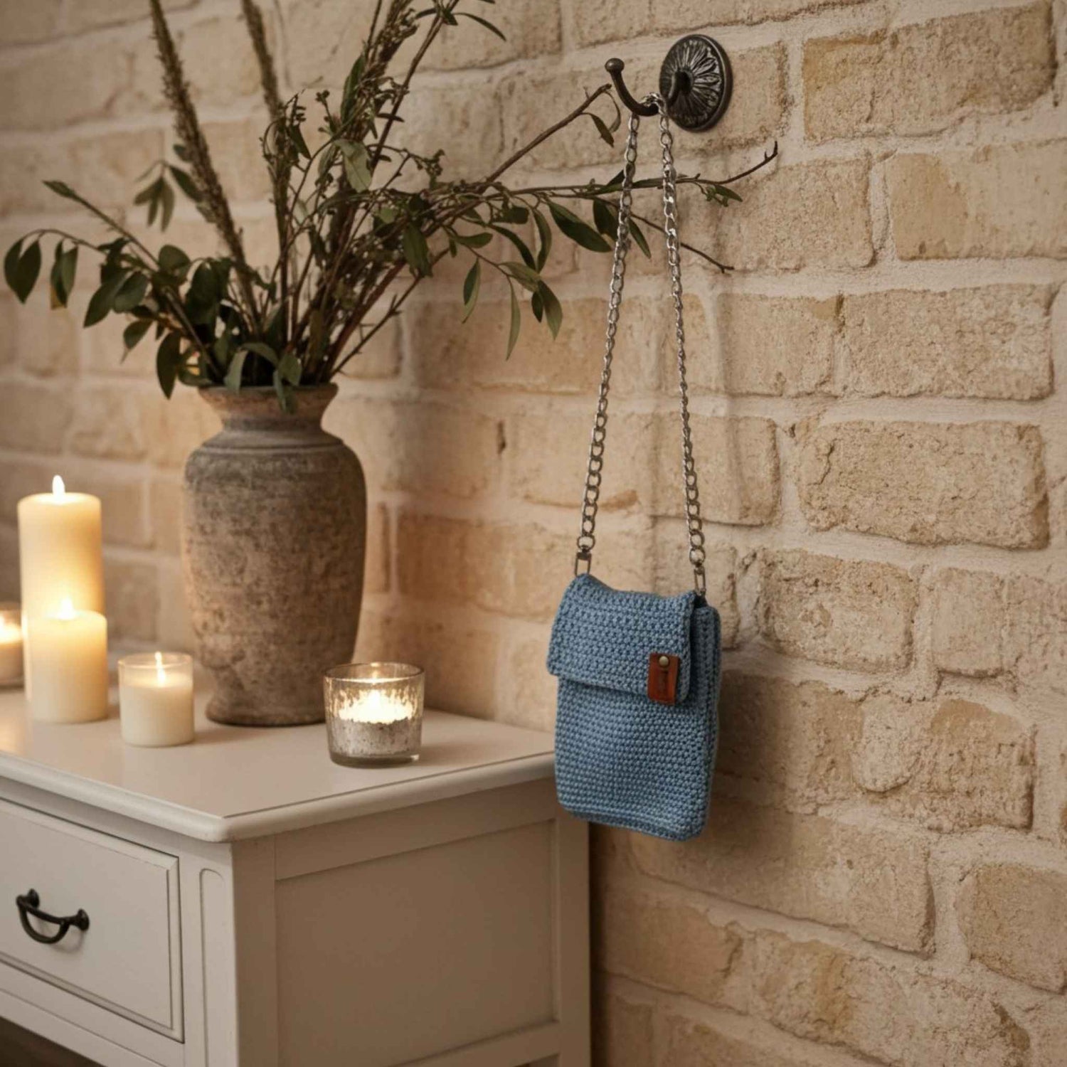 Blue knitted bag hanging on a brick wall with candles and a vase in the background.