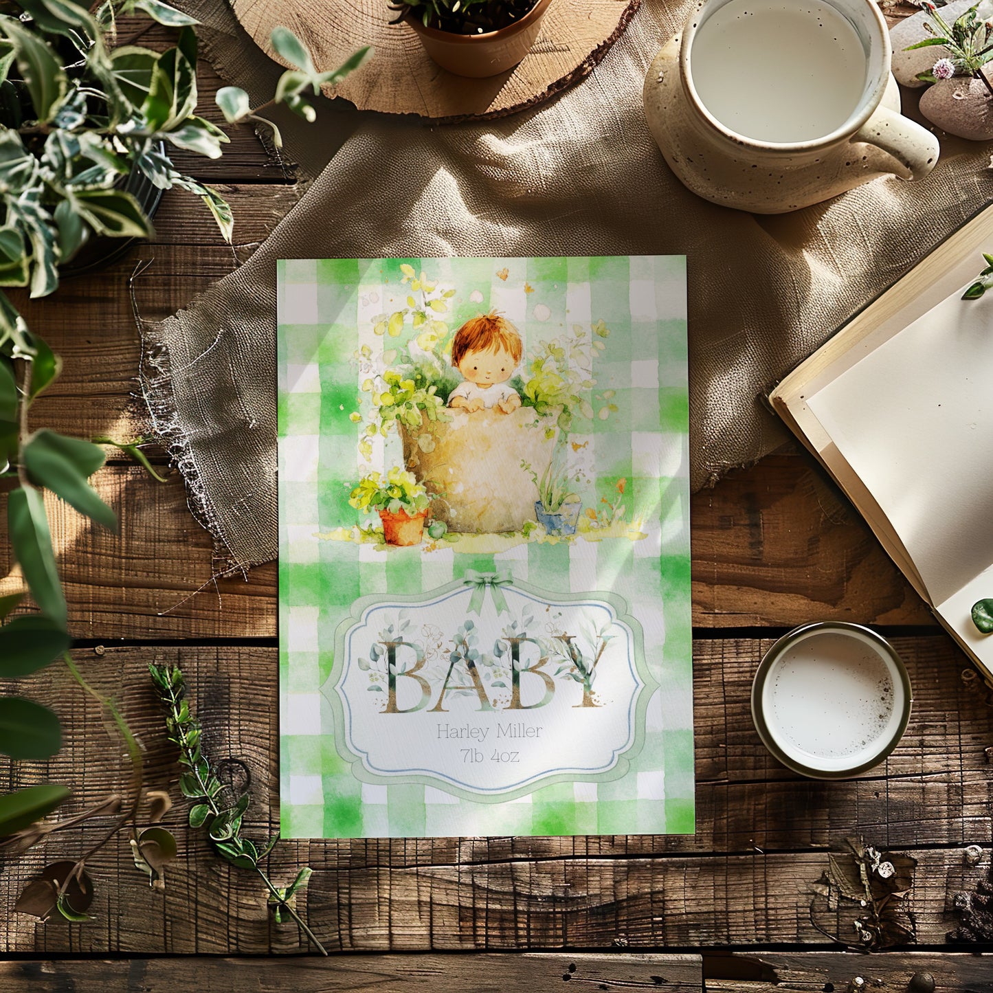 A personalised baby card with a green and white gingham pattern and a vintage-style illustration of a baby in a garden pot, styled on a rustic wooden table with a mug, book, and plants.