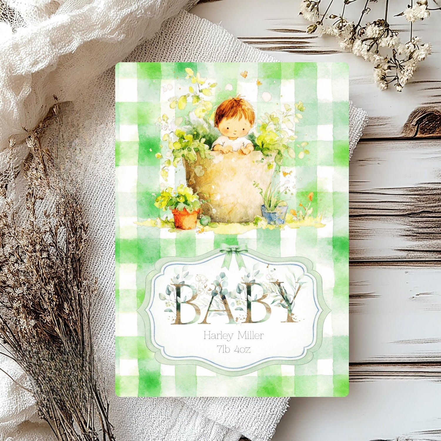 A baby shower card with a green and white gingham border, featuring a watercolour illustration of a baby in a flower pot surrounded by flowers, and the word 'BABY' in a decorative label. The card is styled on a rustic white wooden surface with dried flowers and a textured cream fabric.