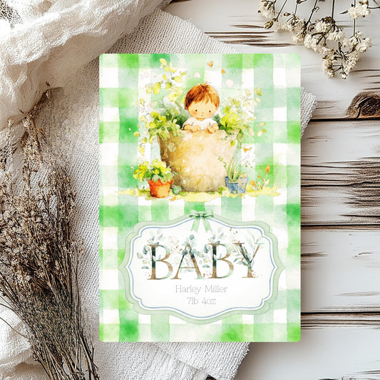 A baby shower card with a green and white gingham border, featuring a watercolour illustration of a baby in a flower pot surrounded by flowers, and the word 'BABY' in a decorative label. The card is styled on a rustic white wooden surface with dried flowers and a textured cream fabric.