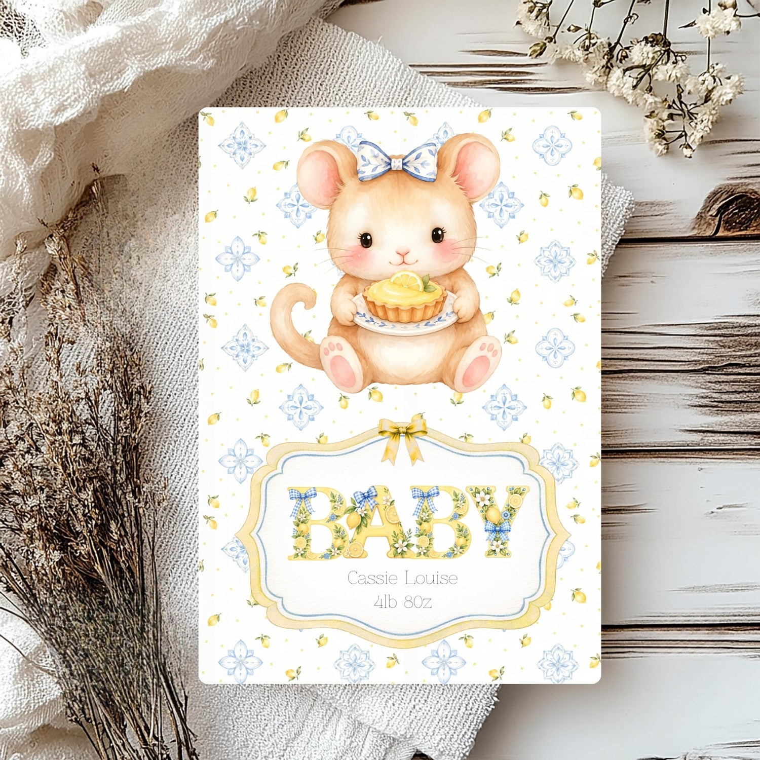 A baby shower card featuring a cute cartoon mouse holding a cupcake, with the word 'BABY' in floral lettering, styled on a rustic wood surface with dried flowers and fabric.