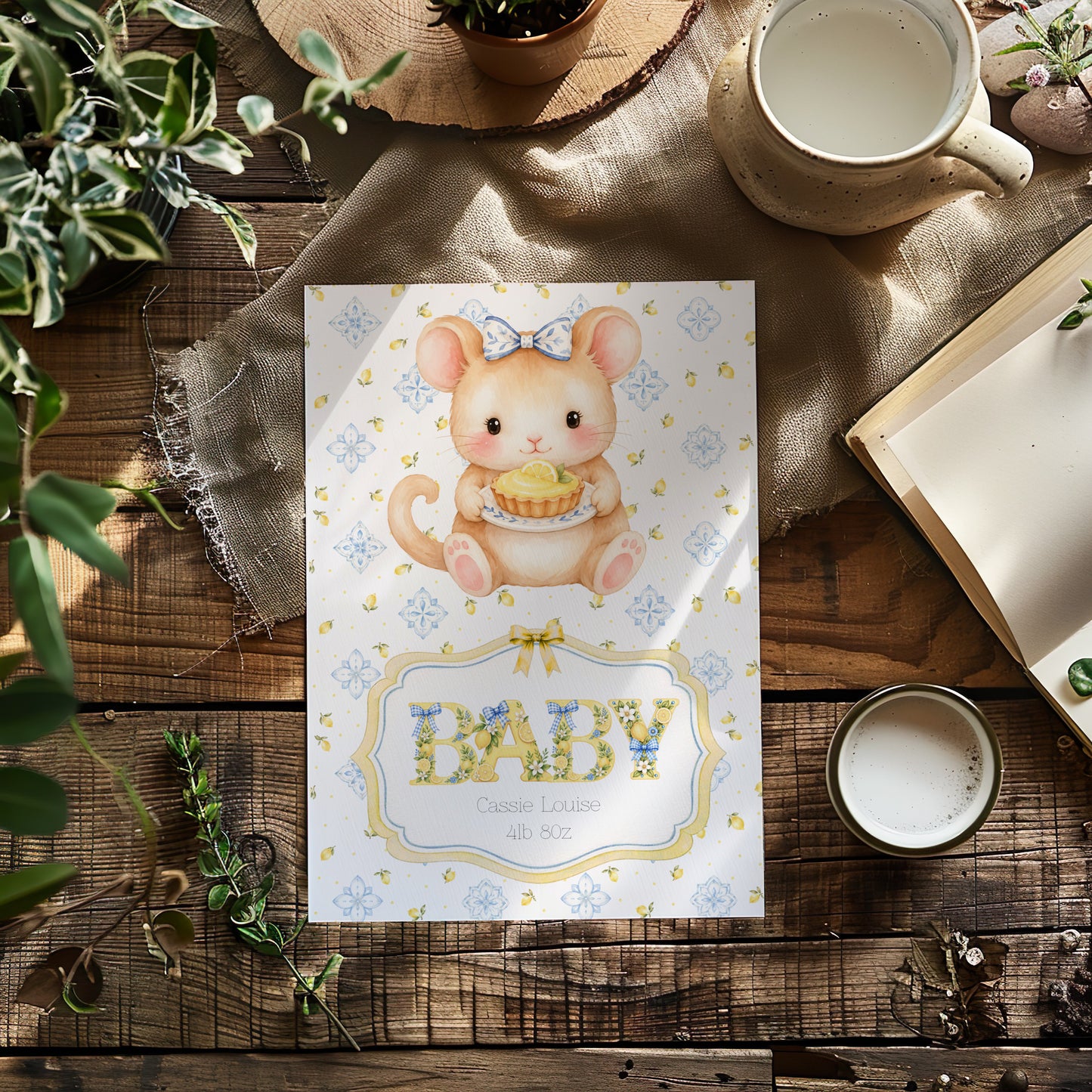 A baby card featuring a cute watercolour illustration of a baby mouse holding a cupcake, with the word 'BABY' in decorative letters. The card is styled in a flat-lay arrangement on a rustic wooden table with a mug, book, and greenery.