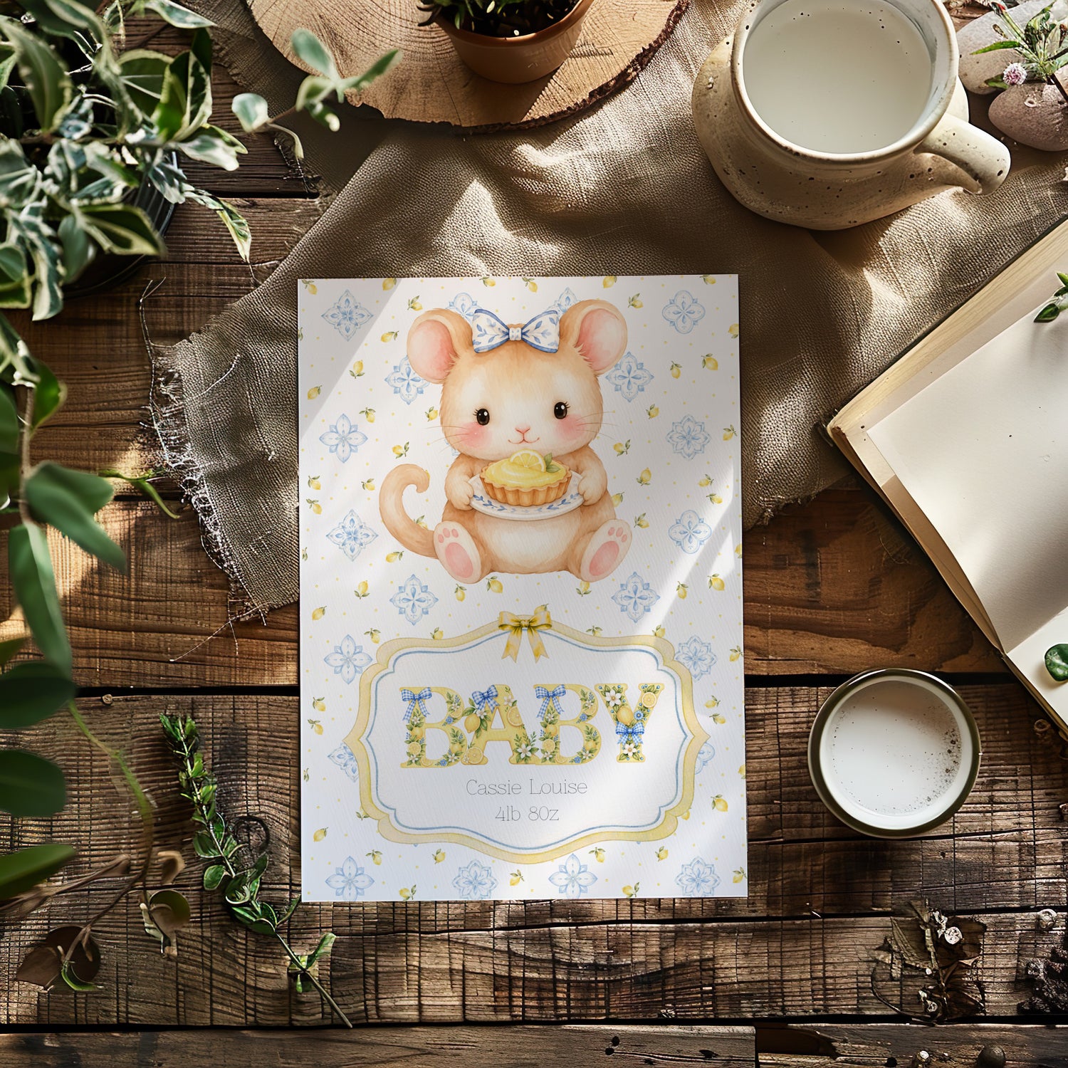 A baby card featuring a cute watercolour illustration of a baby mouse holding a cupcake, with the word 'BABY' in decorative letters. The card is styled in a flat-lay arrangement on a rustic wooden table with a mug, book, and greenery.