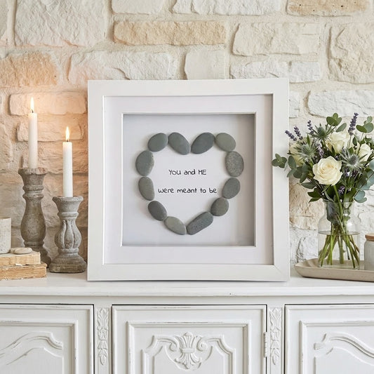 A white square shadow box frame displayed on a white cabinet against a light stone wall. Inside the frame, smooth grey stones are arranged in a heart shape around the words 'You and ME we're meant to be' in black script.