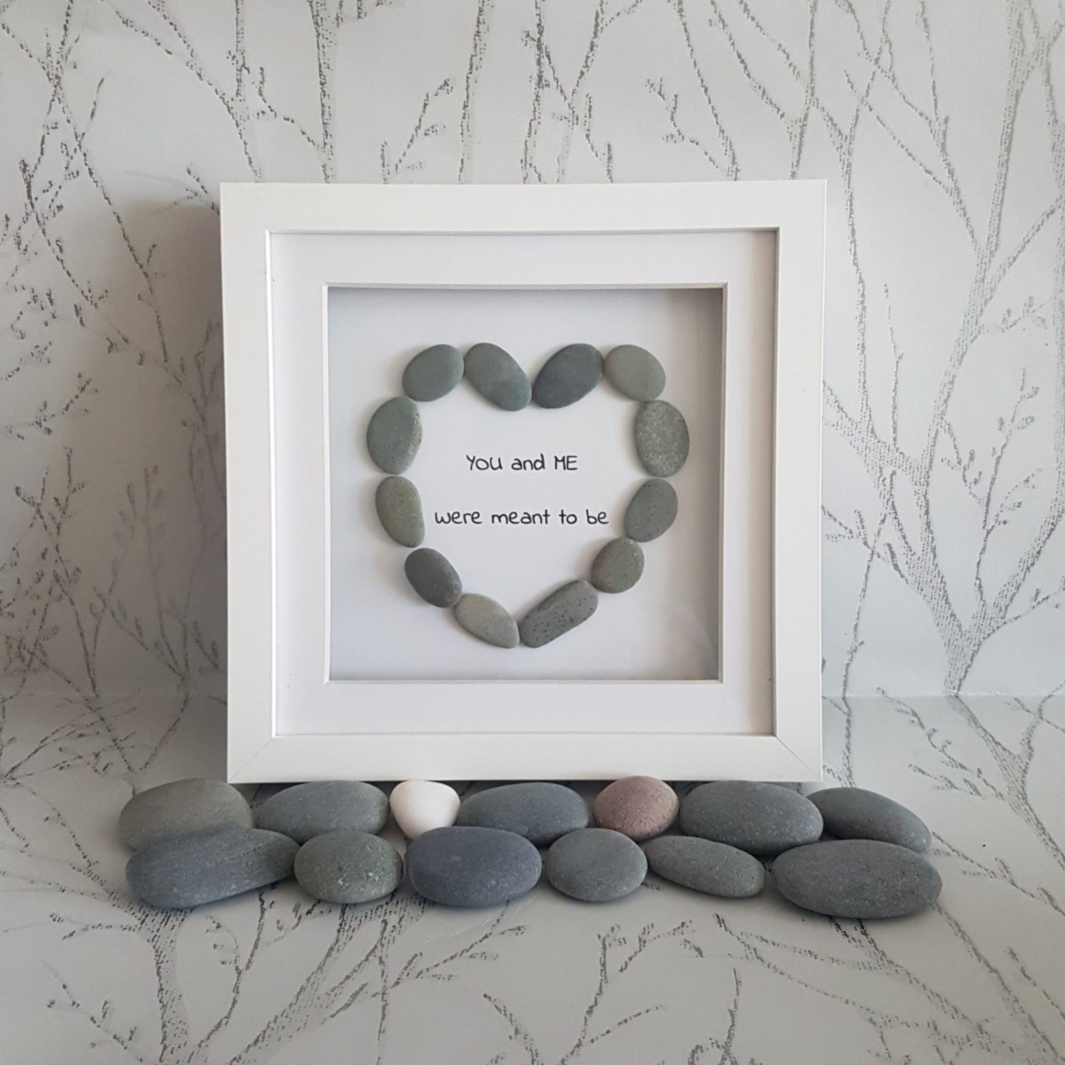 A heart-shaped pebble art picture frame with the inscription 'You and me were meant to be' positioned in a white frame, surrounded by various coloured pebbles.