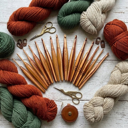 A flat lay of wooden crochet hooks arranged in a fan shape, surrounded by colourful yarn skeins in rust orange, sage green, cream, and natural beige, with two metal scissors and a small brown leather case on a whitewashed wood background.