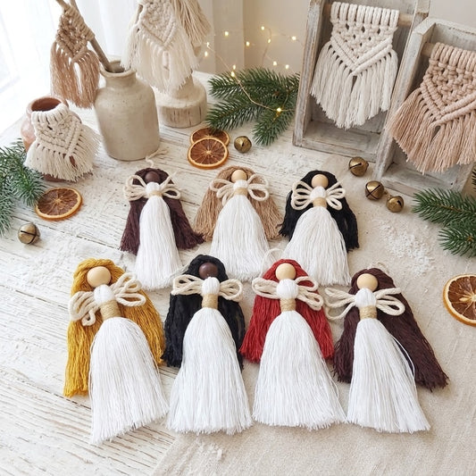 A set of handmade tassel angels arranged on a light fabric surface with boho-style props including a ceramic vase, greenery, dried orange slices, and macrame wall hanging decor.
