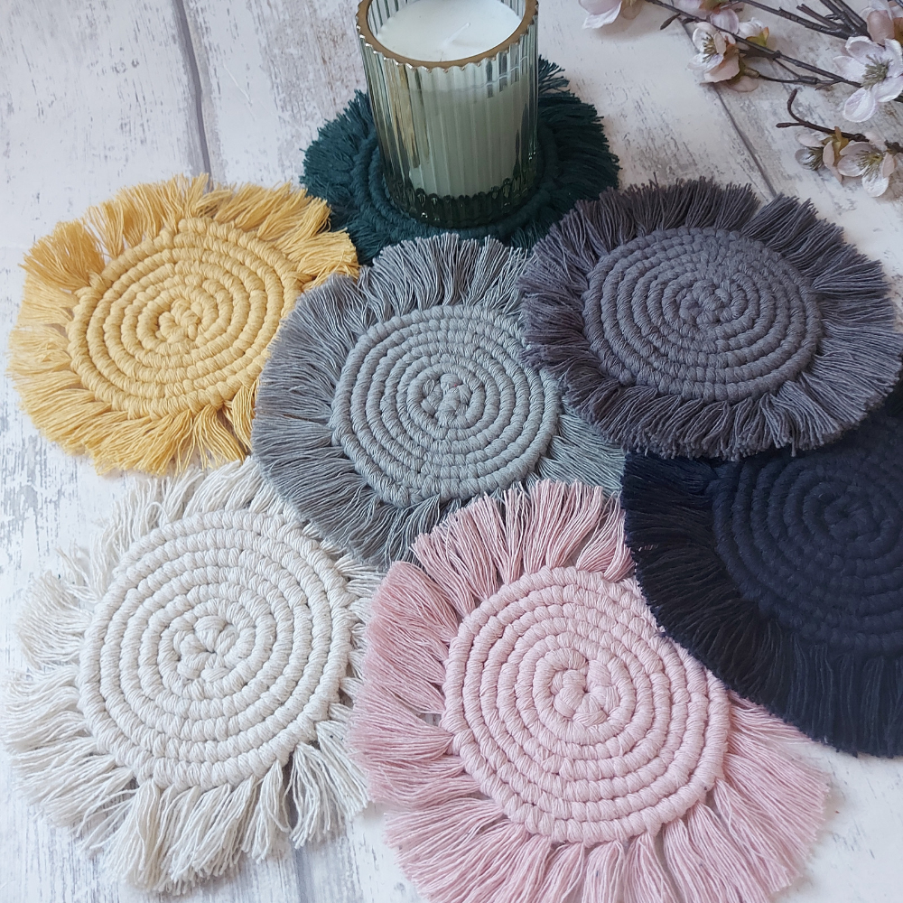 A collection of round macrame coasters in various colours displayed on a white surface, with a cup and some flowers in the background.