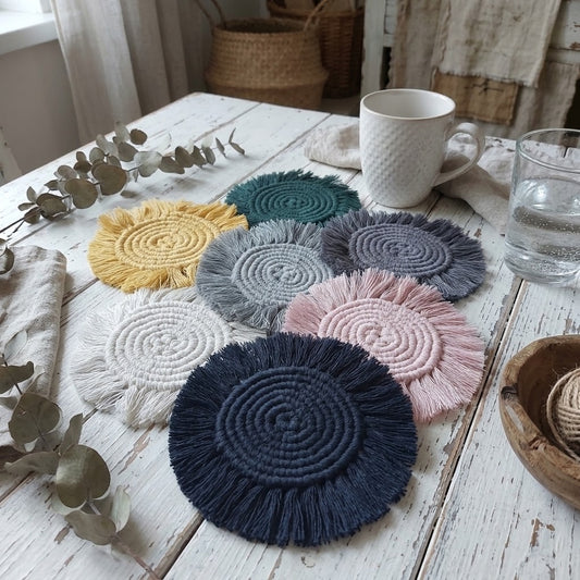 Six round fringed woven coasters in assorted colors including navy blue, pink, light grey, teal, yellow, and lavender, arranged on a rustic white wooden table with a mug, glass of water, and eucalyptus branches.