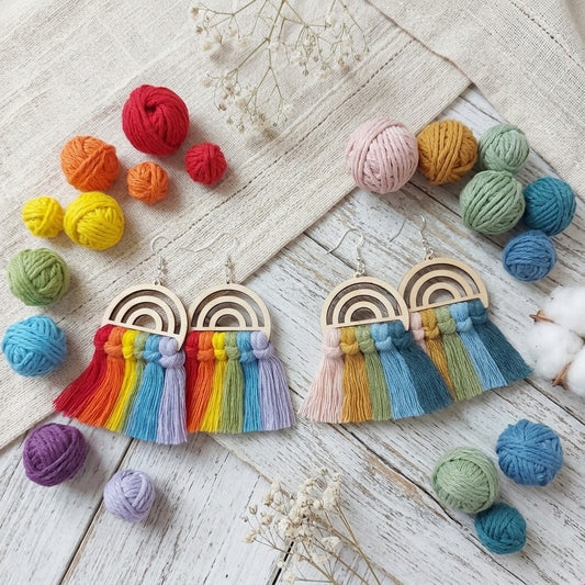 A pair of handmade rainbow tassel earrings displayed on a rustic white wooden surface with colourful yarn balls and dried flowers. The earrings feature wooden arches with layered rainbow-colored tassels in red, orange, yellow, green, blue, and purple.